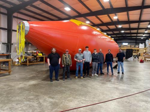 Crew standing next to large orange "Seaslug" towable bladder in warehouse